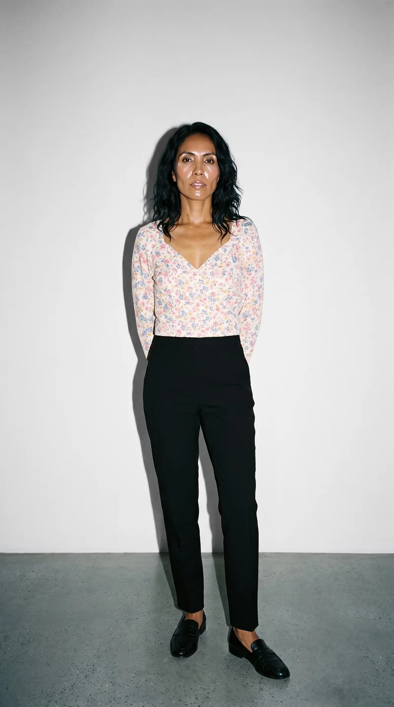 A 45-year-old Hispanic woman wears a floral print wrap top in a full-body front view. She is smiling with her hands behind her back against a Fog Grey studio-wall backdrop.