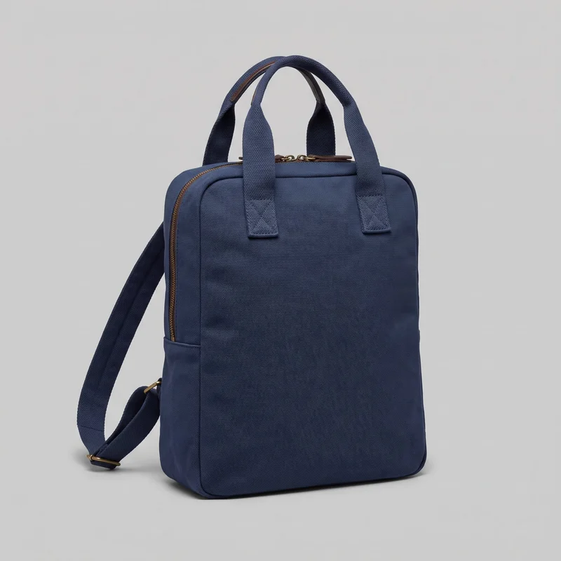 A navy canvas backpack tote is shown as a mostly complete standalone product view, angled to reveal its flat front panel and one side. The structured rectangular bag has softly rounded corners, a squared base, two short arched top handles, exposed adjustable backpack straps, and a zipper running around the top edge and down the side.