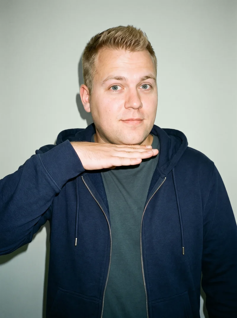 A 33-year-old Caucasian man is shown in a bust-length front three-quarter view wearing a men's navy zip-up hoodie. One hand is raised to his jaw, and he has a curious expression. A sage green studio-wall backdrop fills the background.