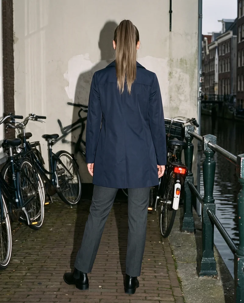 A 23-year-old Caucasian woman stands in a relaxed pose, shown full body from the back while wearing a women's navy trench coat. A canal bridge with bicycles forms the backdrop.
