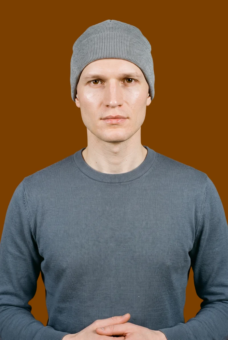 A 38-year-old Slavic man is shown from the front in an upper-body view wearing a grey kufi. His hands are held in front, and he has a neutral facial expression. The image has a flat chocolate-brown backdrop.