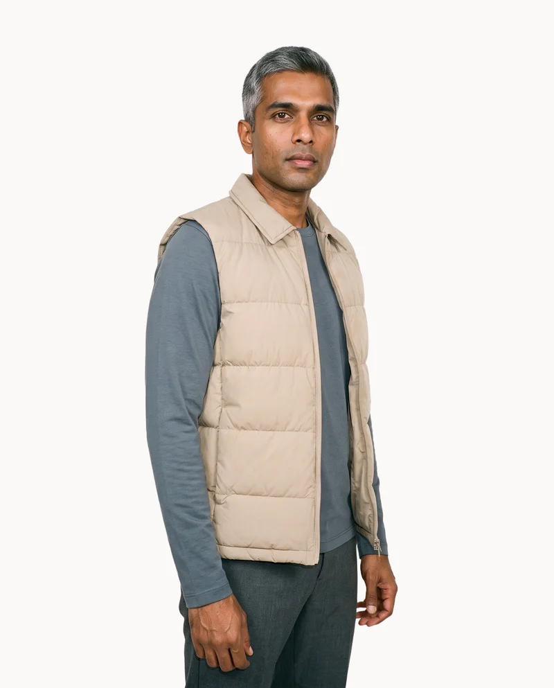 A 37-year-old Indian man wears a beige down gilet in a relaxed standing pose, shown from the front at a three-quarter angle in a three-quarter-length frame. His serious expression is visible against a flat Chantilly Lace-colored backdrop.
