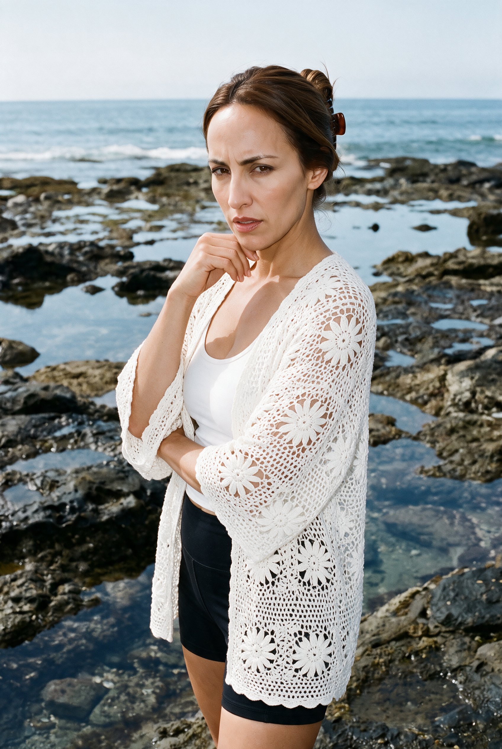 A 35-year-old Hispanic woman wears a white crochet cover-up, shown from the upper body at an angled front view with her wrist raised to her jaw and an intense expression. Tidal pool rocks form the backdrop.