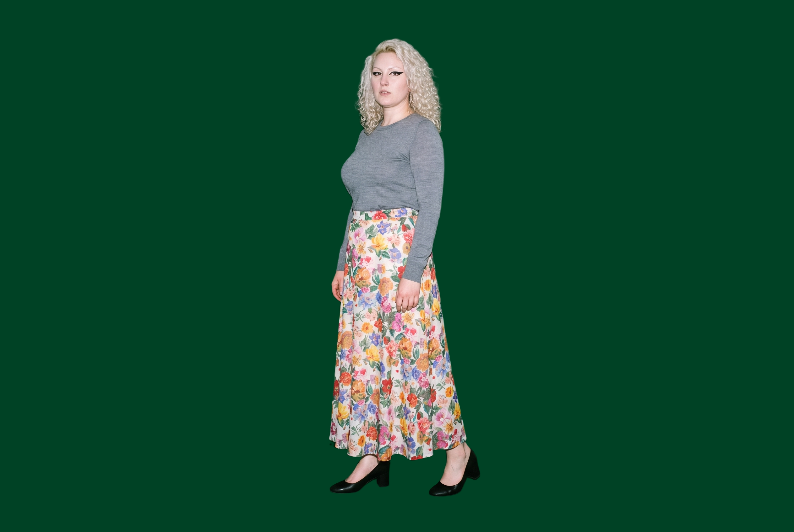 A 32-year-old Caucasian woman wears a floral print maxi skirt in a full-body view from a front three-quarter angle. Her ankles are crossed, and she has a neutral facial expression against a British Racing green flat-color backdrop.