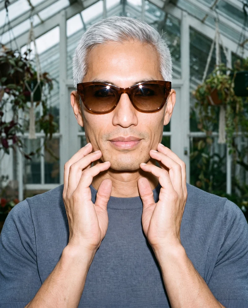 A 46-year-old Southeast Asian man wears brown cat-eye sunglasses in a front-view bust composition, with his hands positioned at his jaw and a confident expression. A greenhouse conservatory serves as the backdrop.