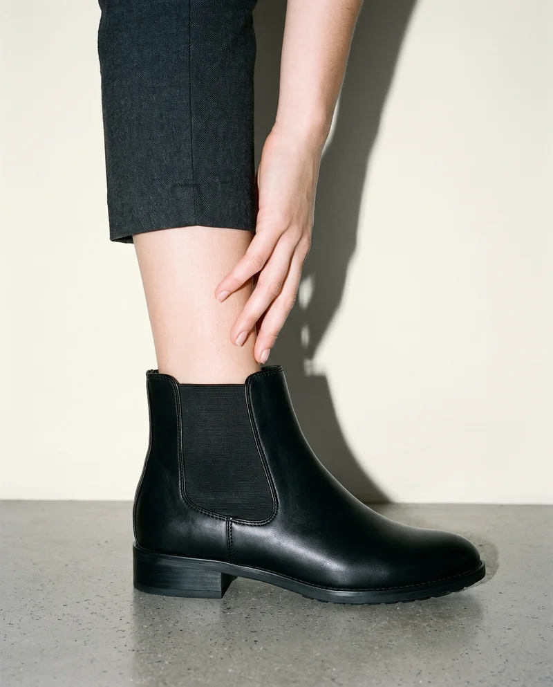 A woman’s black leather Chelsea boot is shown in side view with a hand at the ankle. The ankle-framed product is set against an alabaster studio-wall backdrop.