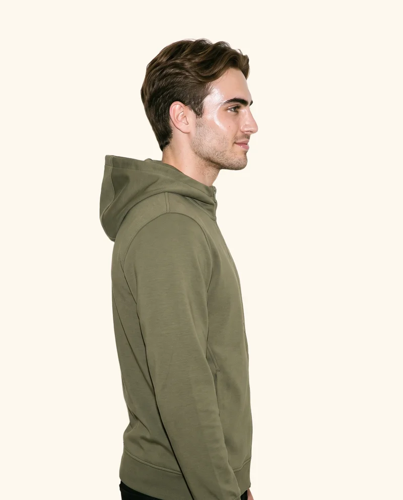An 18-year-old Caucasian man is shown from the upper body in side view, wearing a men's olive zip-up hoodie. He has a shoulder-drop pose and a smiling expression against a creamy white backdrop.