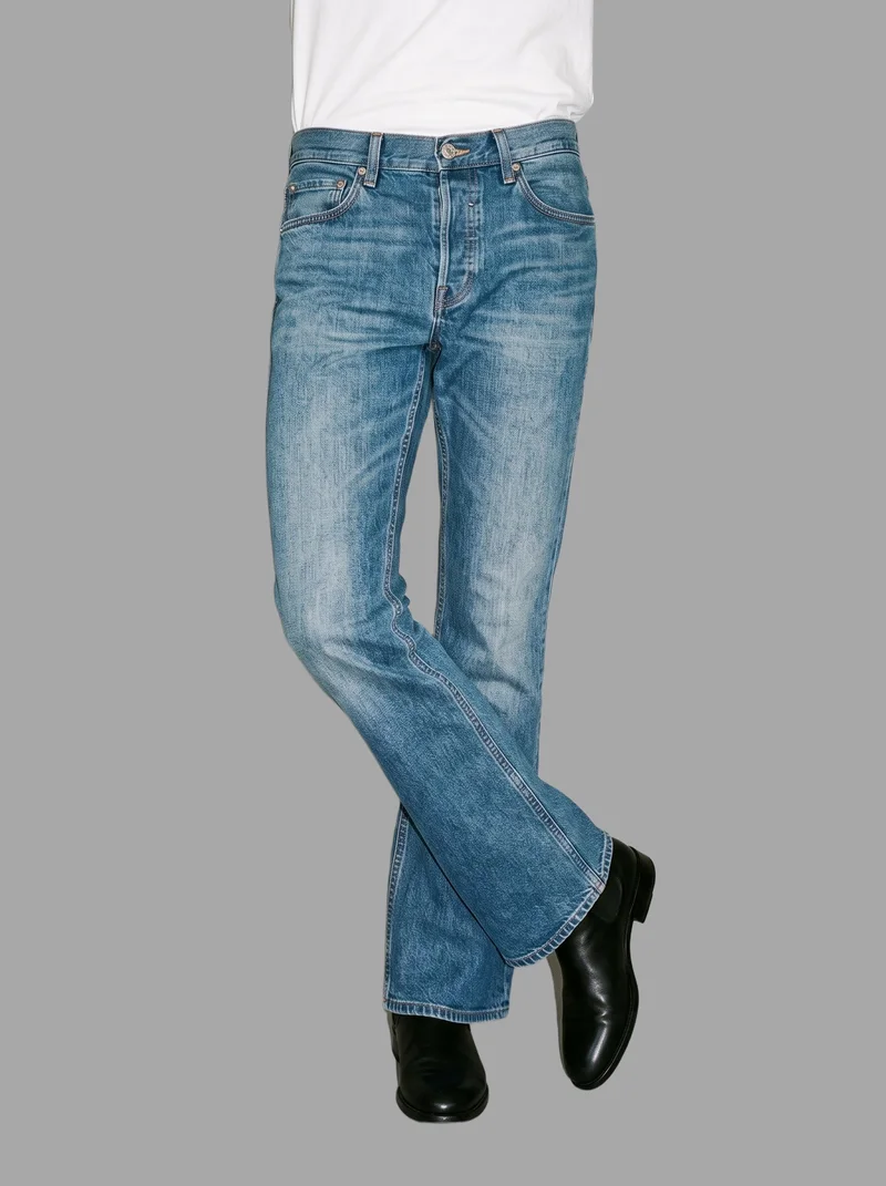 A male model is shown from the lower body in a front view wearing men's mid-wash bootcut jeans, with his ankles crossed. The jeans are presented against a flat pebble grey backdrop.