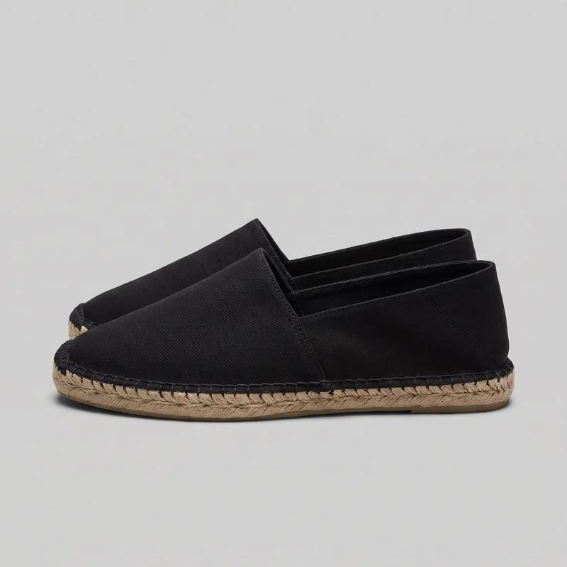 A pair of black low-profile slip-on espadrilles is shown from a side three-quarter angle. The shoes have closed rounded toes, low-cut openings, visible side elastic gussets, stitched side and heel panels, bound toplines, flat heels, and rope-wrapped midsoles with dense whipstitching around the upper edge.