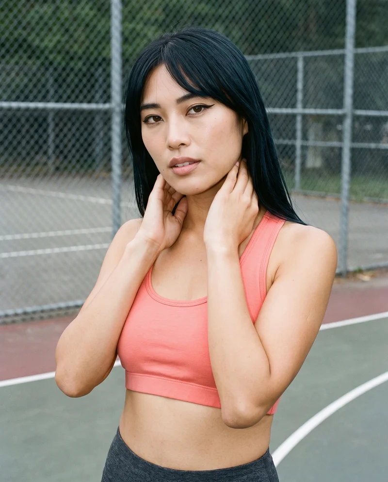 A 28-year-old Japanese woman wears a coral racerback sports bra in an upper-body view. She is shown from a front three-quarter angle with her hands on her neck and a confident expression. A basketball court serves as the backdrop.