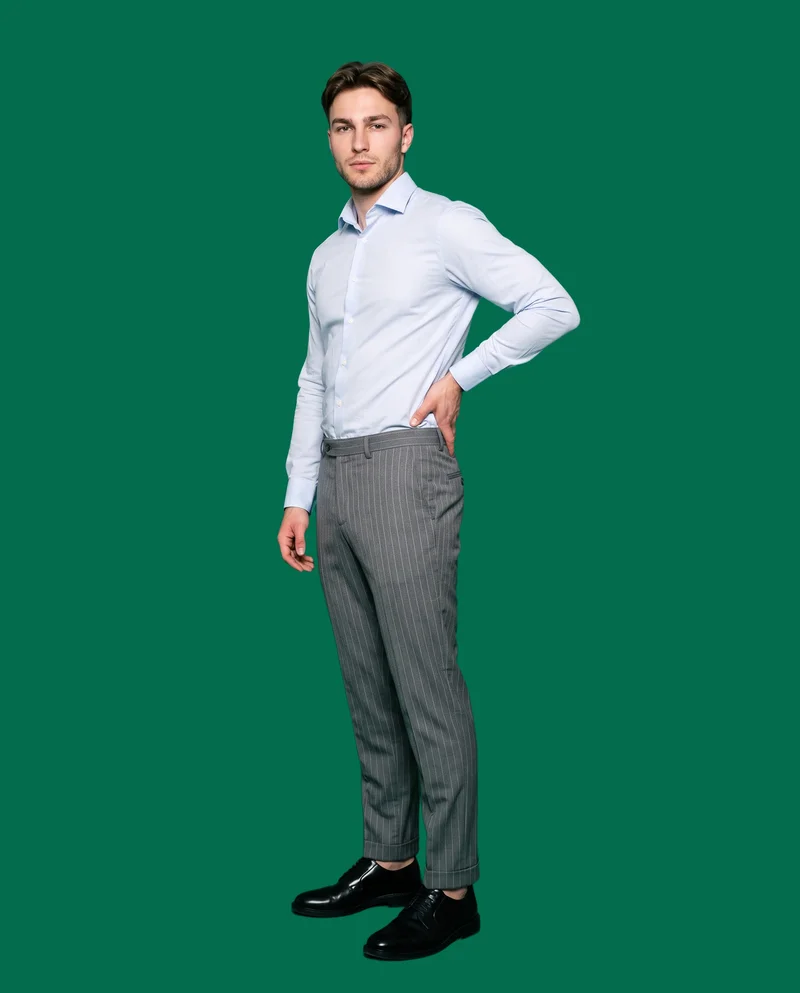 A 24-year-old Caucasian male model is shown full body from a front three-quarter angle wearing grey pinstripe tailored trousers, with one hand on his lower back. The model has a confident facial expression, and the image uses an emerald jewel flat-color backdrop.