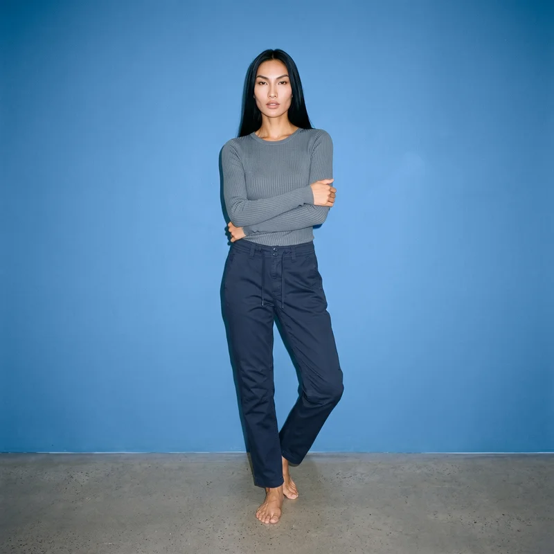 A 32-year-old Southeast Asian woman models navy hiking trousers in a full-body front view. She stands in a self-hug pose with a neutral expression against a steel blue studio-wall backdrop.