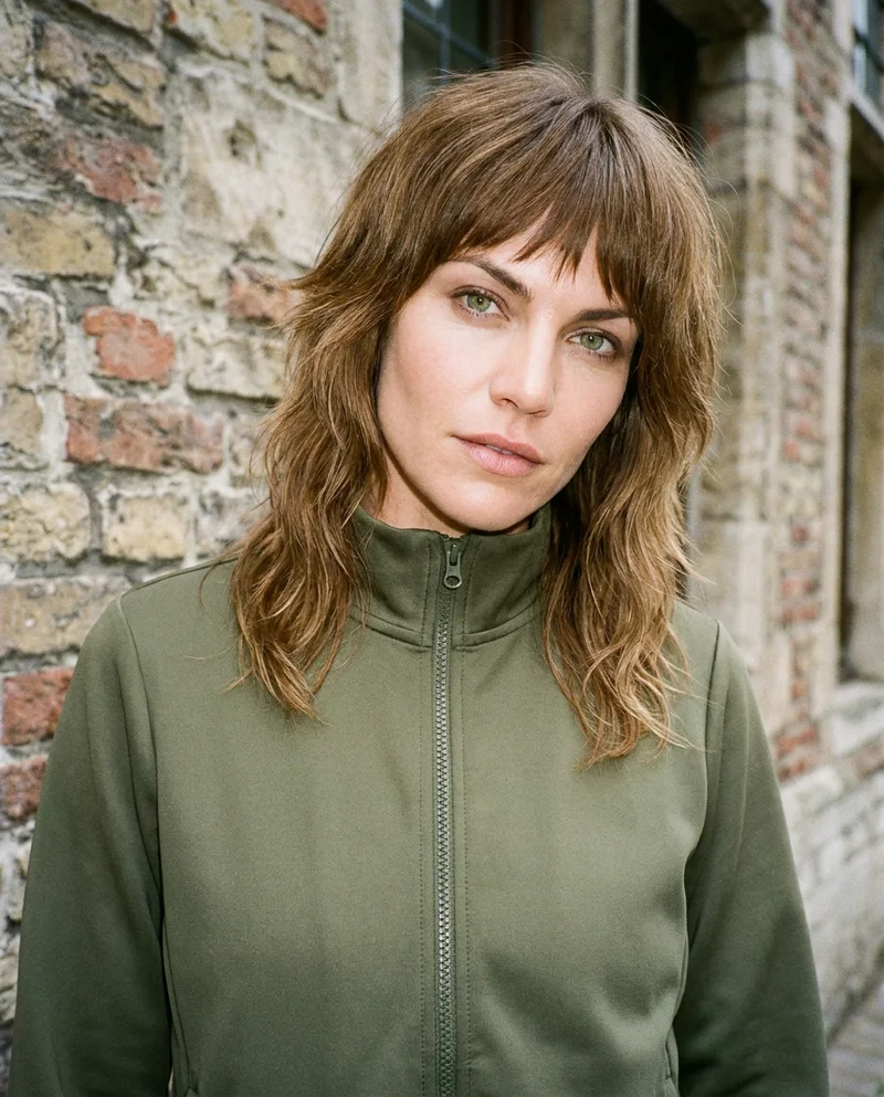 A 36-year-old Caucasian woman wears a women's olive tracksuit in a bust-length frame. She is shown from a front three-quarter angle with her head tilted and a confident expression, against a Belgian cobblestone street backdrop.