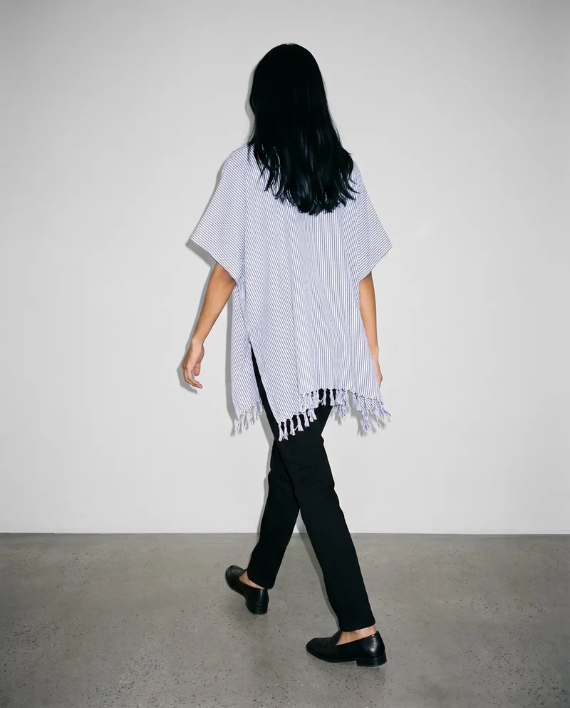 A 24-year-old Indian woman is shown from behind in a full-body view while walking, wearing a women's striped beach cover-up. The Fog Grey studio-wall backdrop provides a plain background.