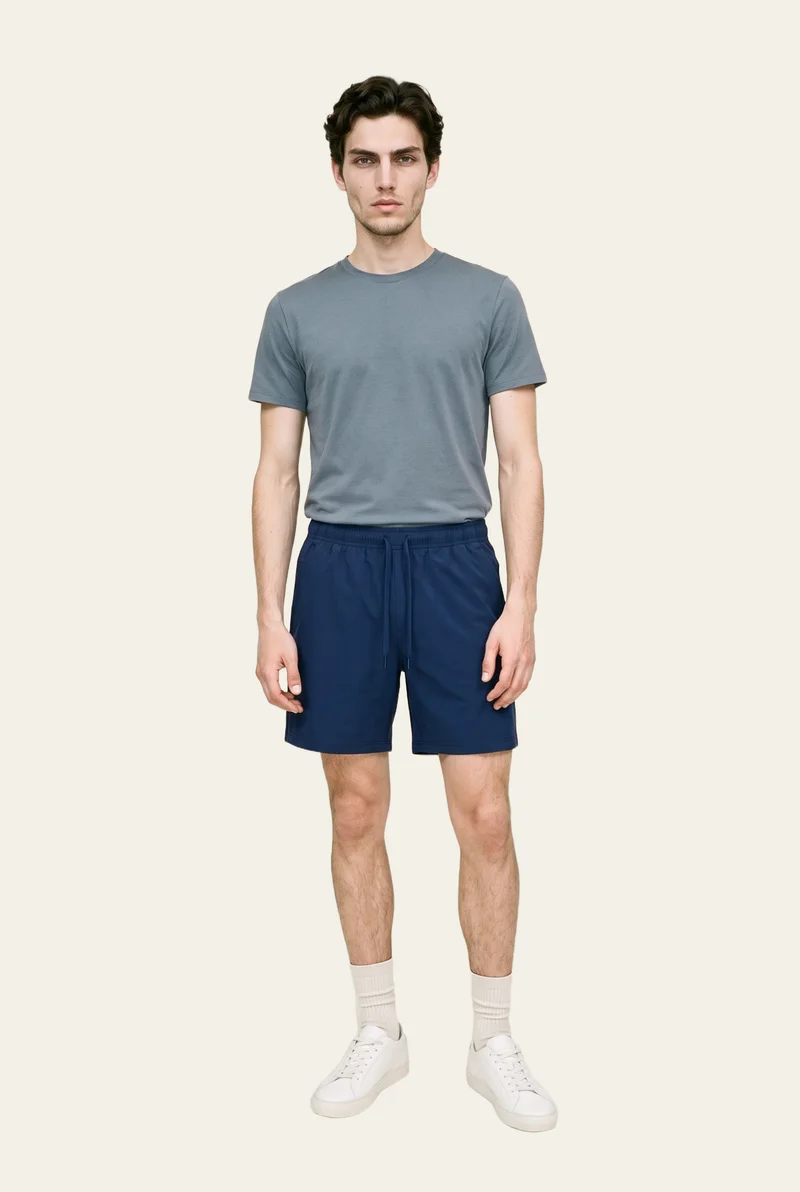 An 18-year-old Caucasian male model wears men's navy running shorts in a full-body front view, standing with his hands in front and a natural facial expression. The model is shown against a flat alabaster backdrop.