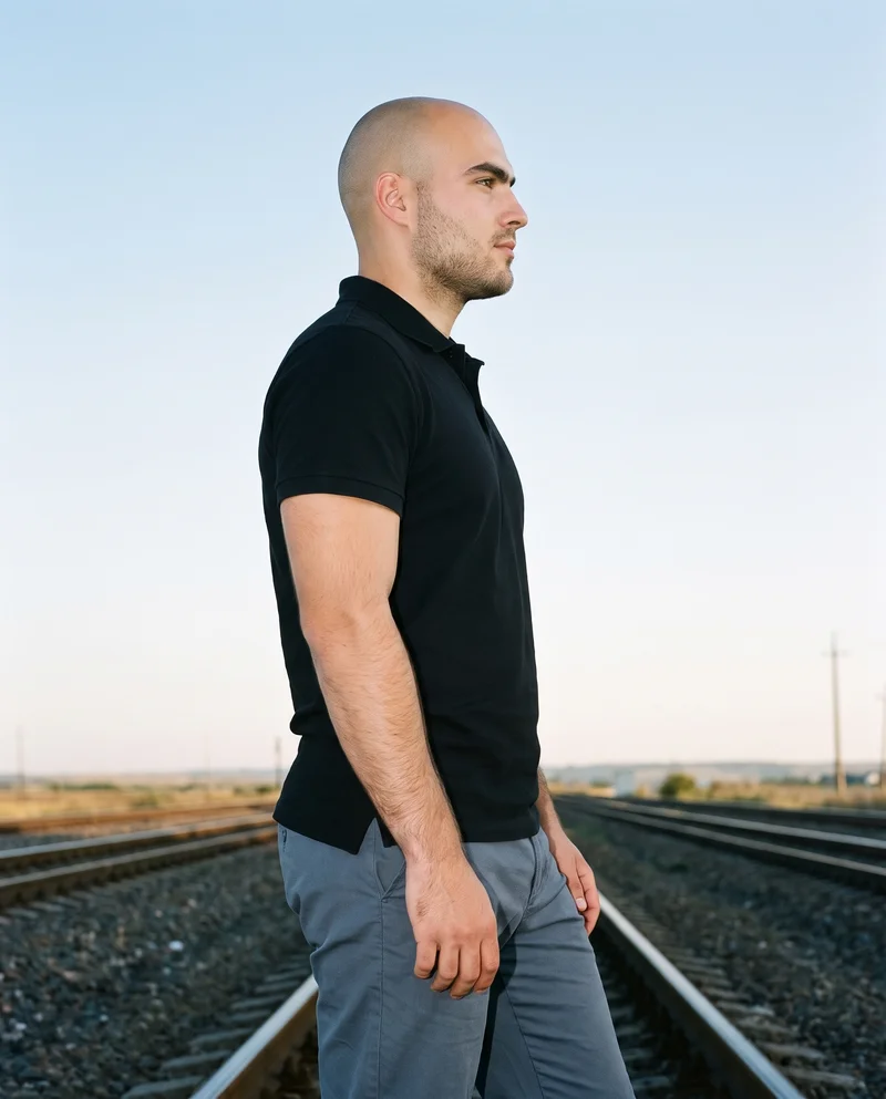 A 23-year-old Caucasian man wears a black polo shirt in a three-quarter-length side view. He is posed with his weight back and a soft facial expression, with train tracks serving as the backdrop.
