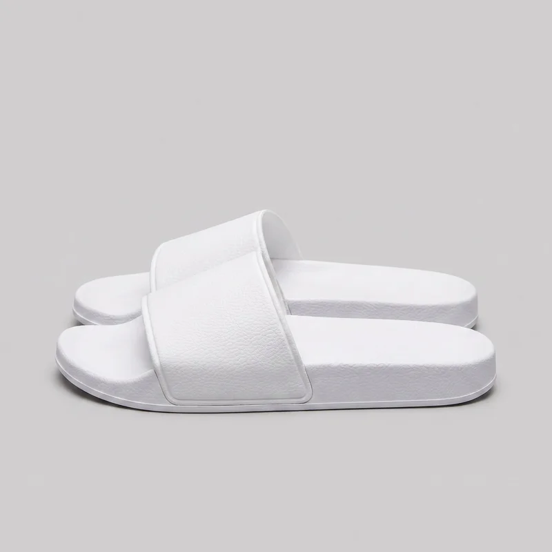 A pair of white slip-on slide sandals is shown in a three-quarter side view, with one sandal positioned in front of the other. Each sandal has an open toe and heel, a broad single strap across the forefoot with a fine pebbled texture and raised edge piping, and a flat platform-style sole with a rounded rectangular outline and raised sidewall.