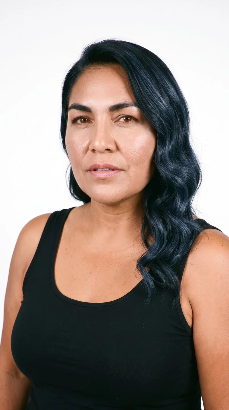A 58-year-old Indigenous American woman with copper-brown skin and a warm golden undertone. Her face has sharp, defined cheekbones and jawlines, with thick eyebrows, brown eyes, and full lips. Her deep black hair is styled in large, polished Hollywood waves.