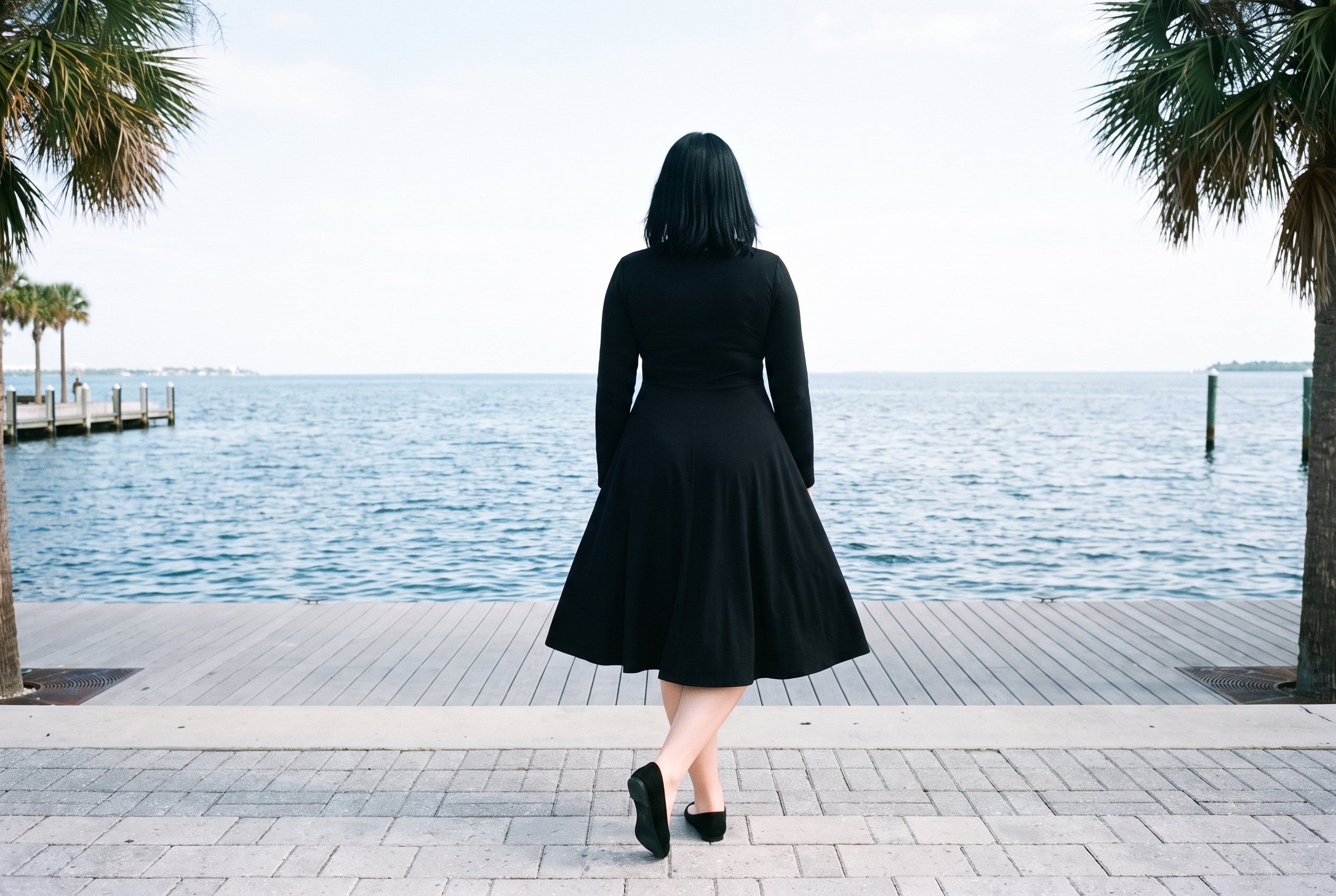 A 29-year-old Slavic woman wears a black modest maxi dress in a full-body rear view, standing with her ankles crossed. Palm trees line a waterfront promenade backdrop.