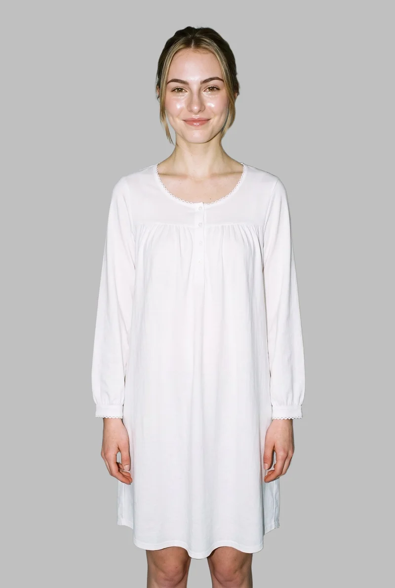 An 18-year-old Caucasian woman stands straight in a front-facing, three-quarter-length view wearing a white cotton nightdress. Her natural expression is visible against a flat silver mist backdrop.