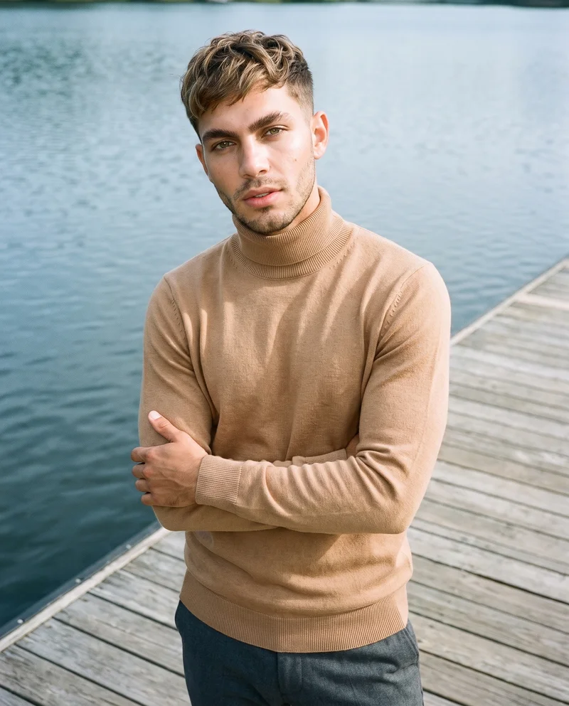A 23-year-old Hispanic man wears a camel roll-neck sweater in an upper-body composition. He is shown from the front at a three-quarter angle with his forearms folded and a natural facial expression. A lakeside dock serves as the backdrop.