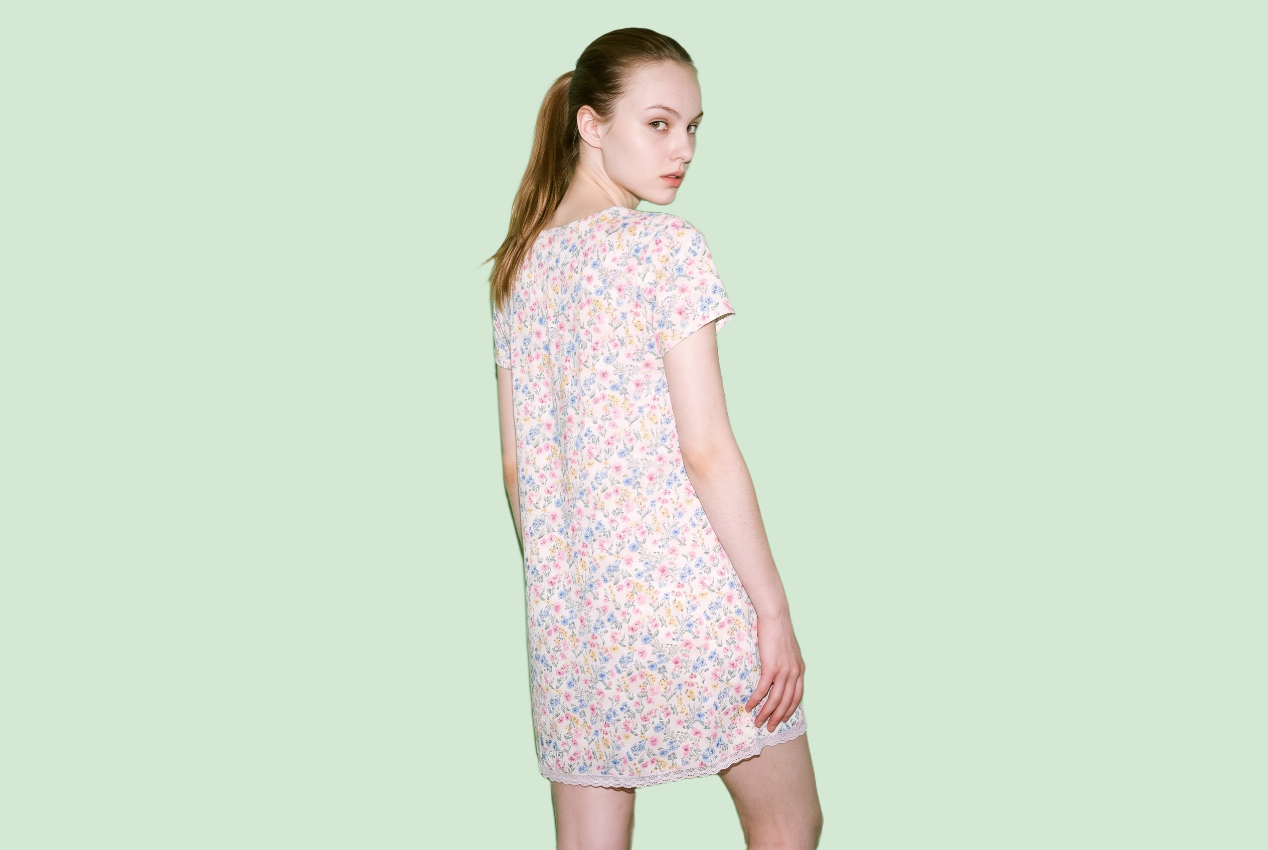 A 21-year-old Caucasian woman wears a floral print nightdress, shown from the back in a three-quarter-length frame with her waist twisted. The garment is presented against a flat pistachio-colored backdrop.
