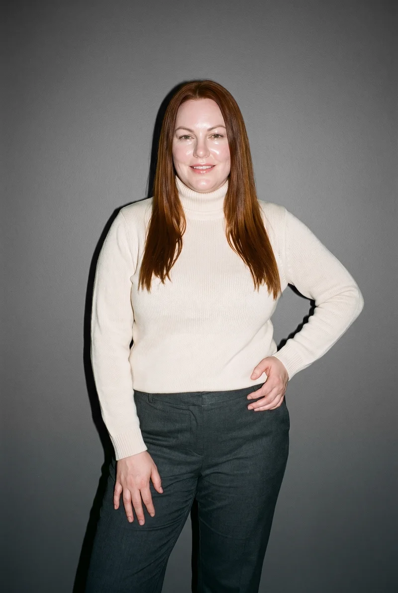 A 37-year-old Caucasian woman wears a cream knit turtleneck, shown from a three-quarter length front view with one hip angled outward and a smile. An Anchor Grey studio-wall backdrop frames the composition.