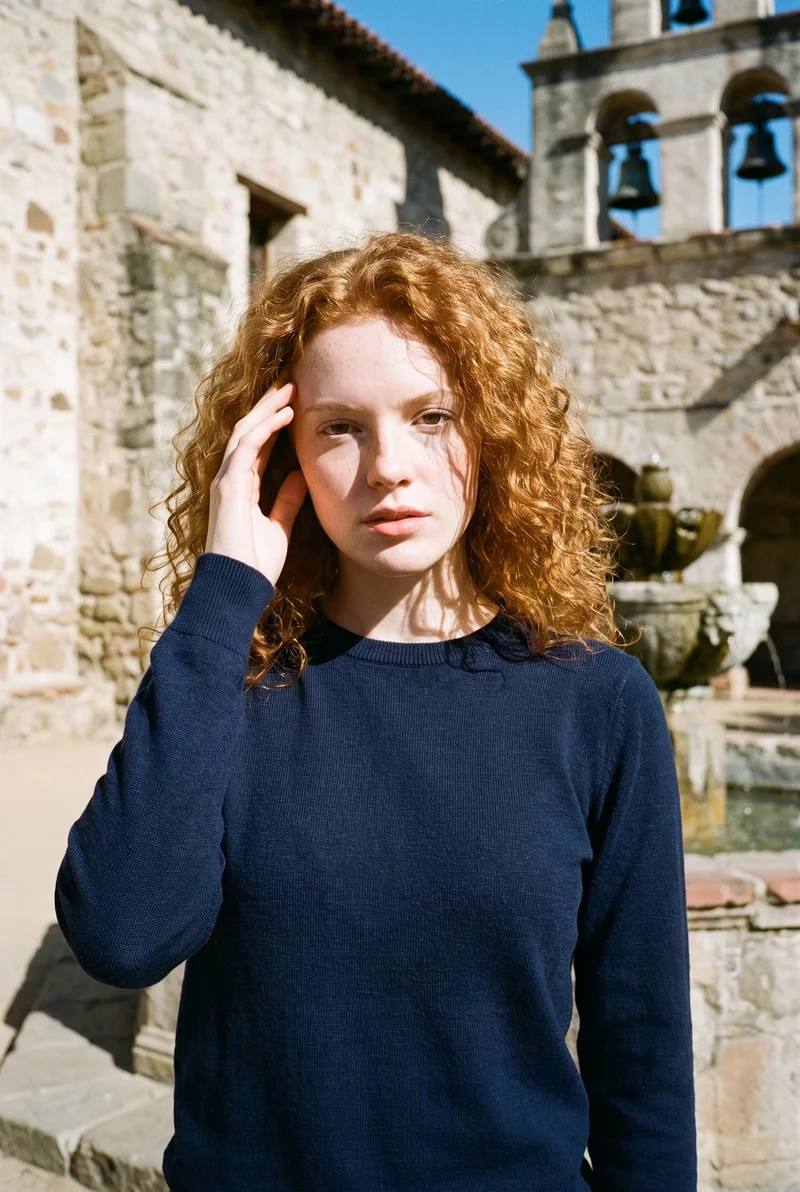 A 22-year-old Caucasian woman wears a navy merino knit, shown from the upper body at a front three-quarter angle with one hand at her temple and a neutral expression. The Mission Courtyard Bell Fountain appears as the backdrop.