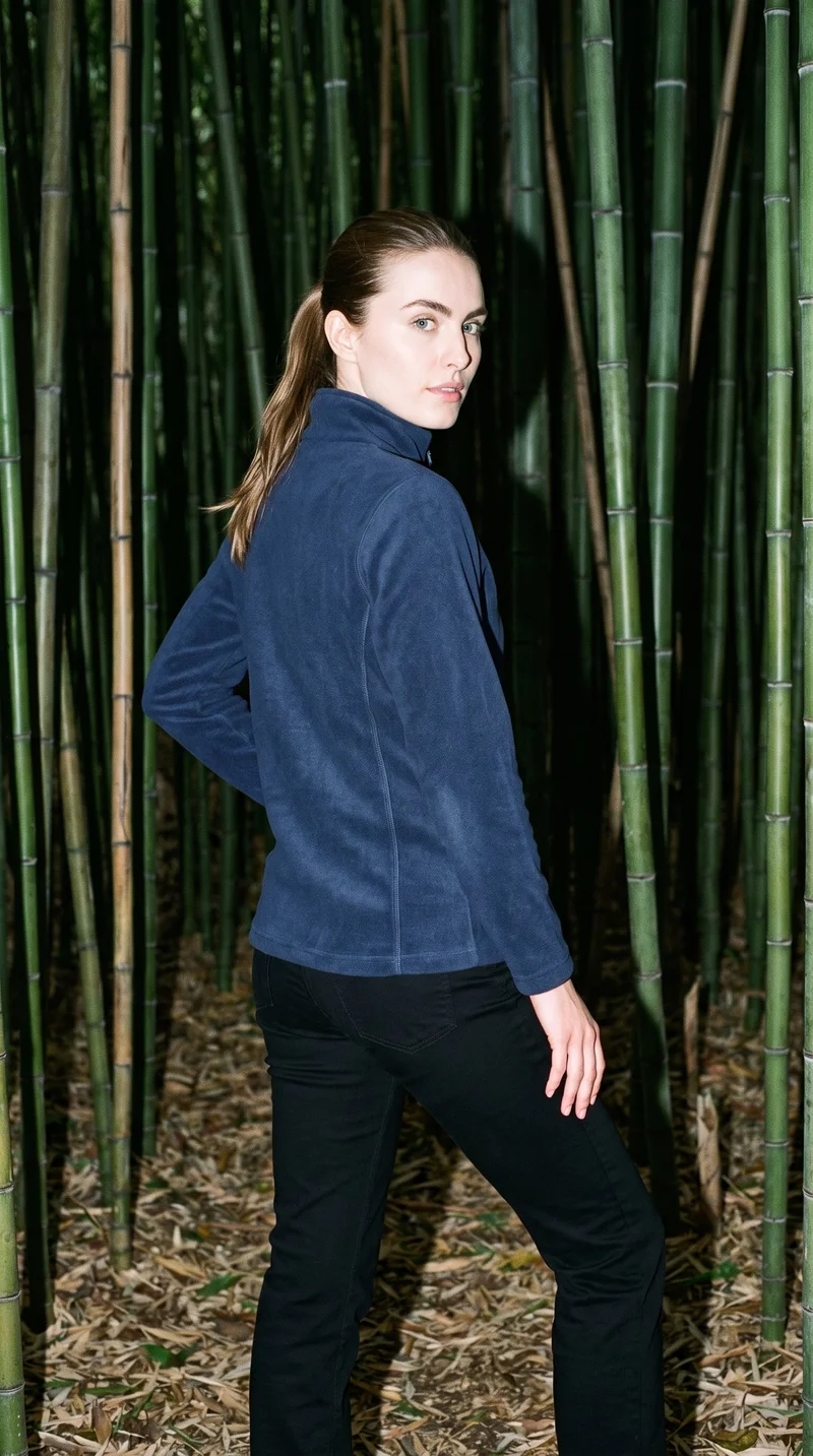 A 29-year-old Caucasian woman wears a women's navy zip-up fleece jacket in a three-quarter-length, angled rear view with one hand on her hip. A bamboo forest forms the backdrop.