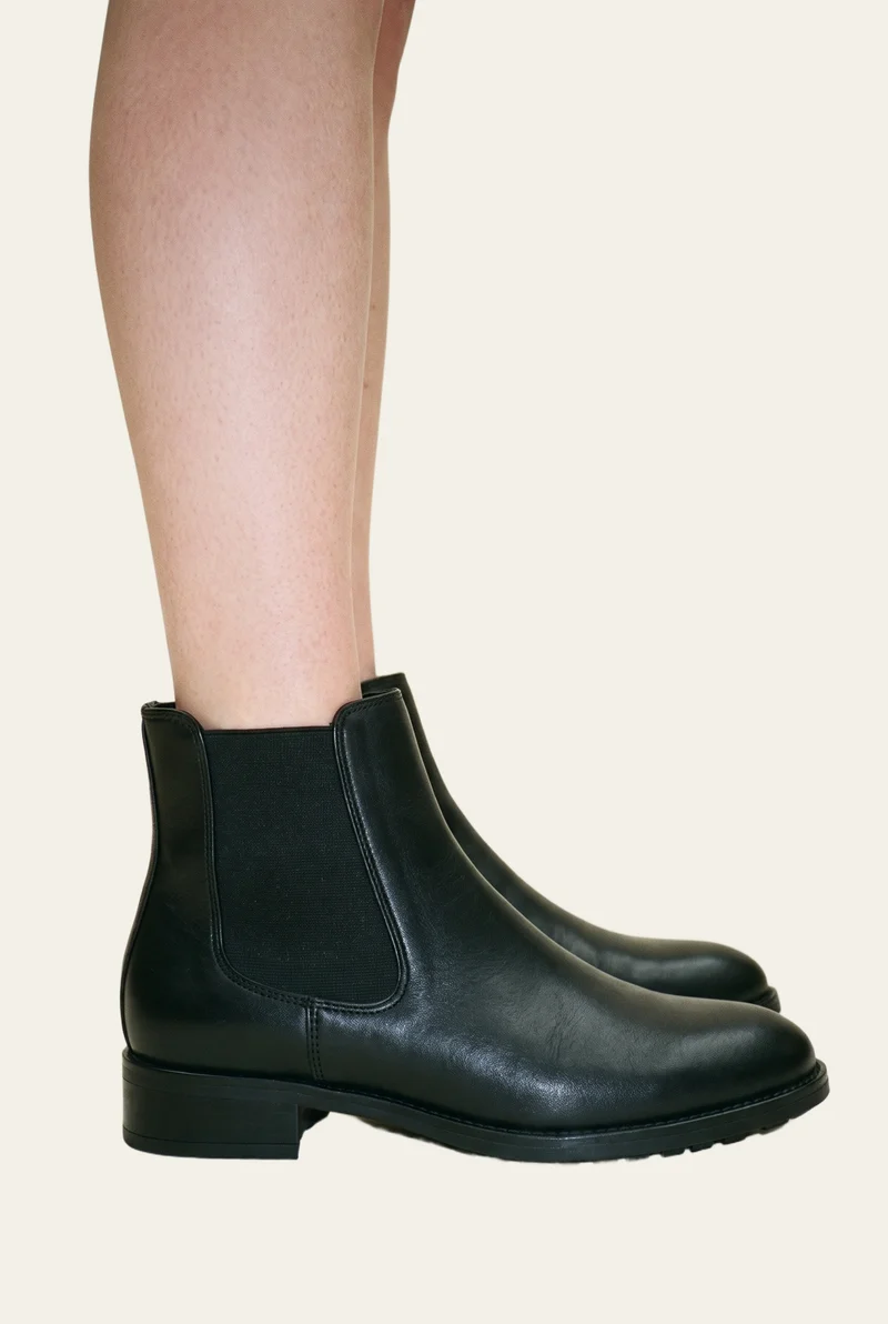 A black leather Chelsea boot is shown from the side with the toe pointed. The ankle-framed boot is set against an alabaster flat-color backdrop.