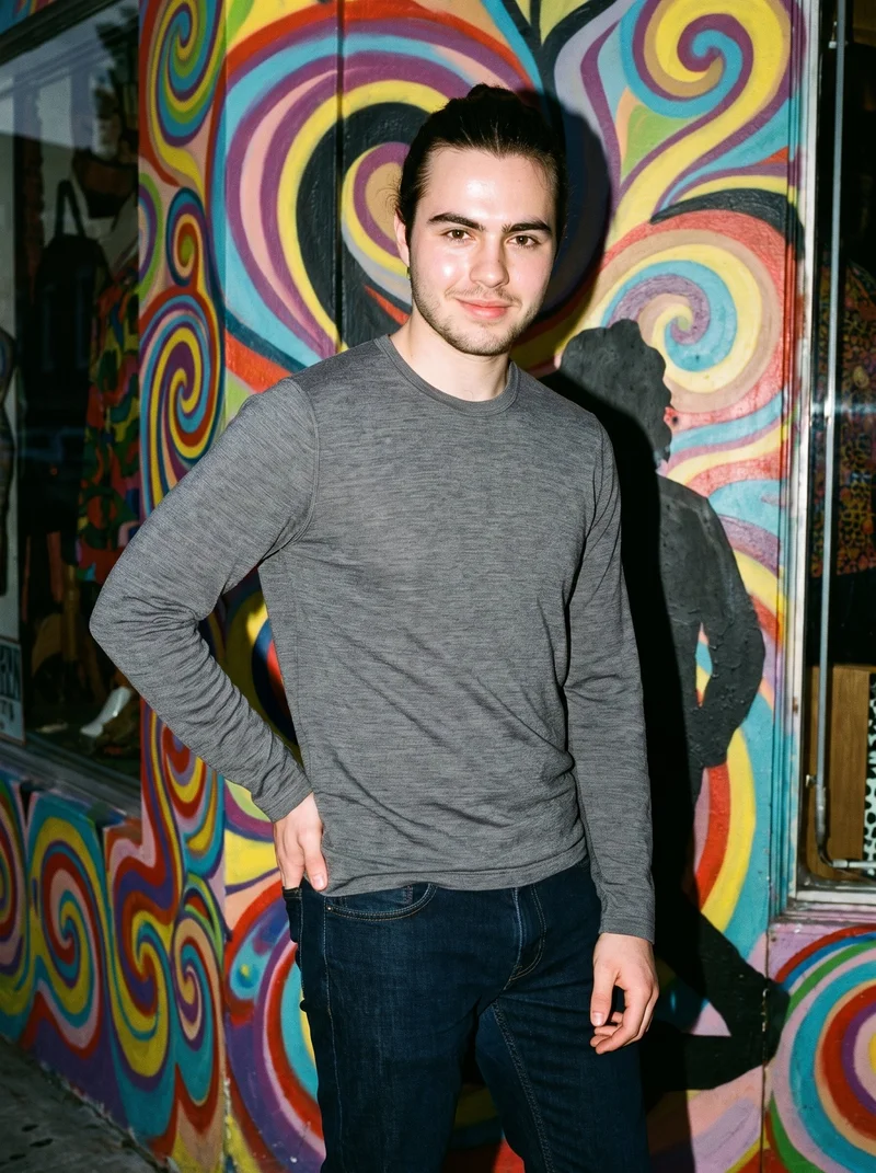 A 19-year-old Caucasian man wears a grey merino thermal top in a three-quarter-length front view. He is smiling with one hand on his hip against a Bohemian District Vintage Psychedelic backdrop.