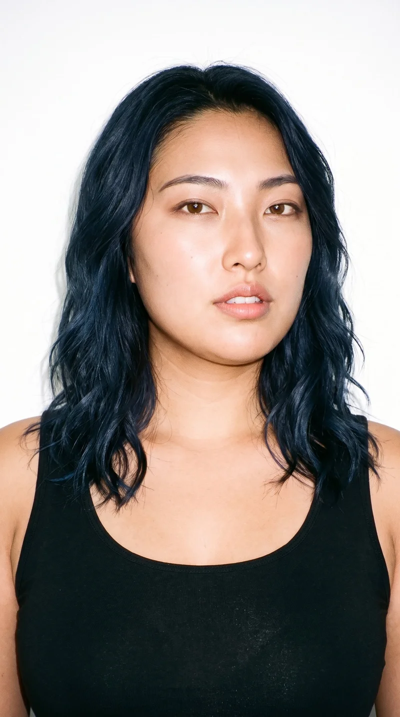 This 25-year-old Japanese woman has light-tan skin with a neutral undertone and East Asian facial features. Her face is oval with balanced proportions and a softly rounded jaw. She has thin eyebrows, very dark near-black eyes, and full lips. Her deep black hair falls in loose, slightly tousled waves with natural volume.