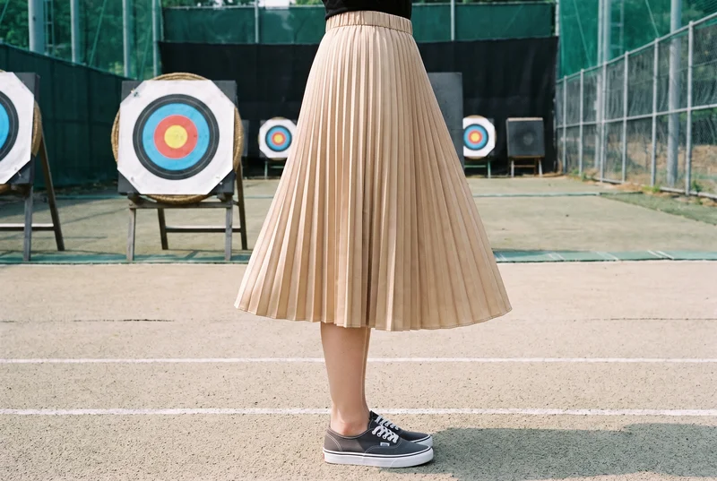 A model wearing a pleated satin midi skirt is shown in a lower-body side view with her feet together. An archery range forms the backdrop.