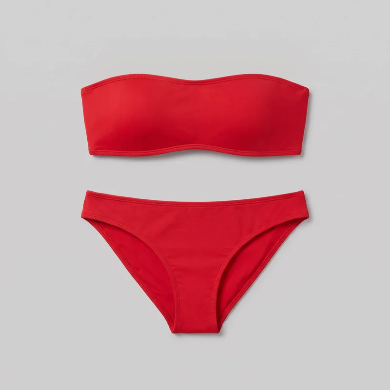 A red two-piece bikini set is arranged flat, with the strapless bandeau top positioned above the matching low-rise brief bottom. The top has a shallow curved upper edge, a gently curved lower edge, straight sides, and visible stitched binding around its edges. The brief has a wide waistband, curved high-cut leg openings, a narrow center-front crotch panel, and stitched binding along the waist and leg edges.
