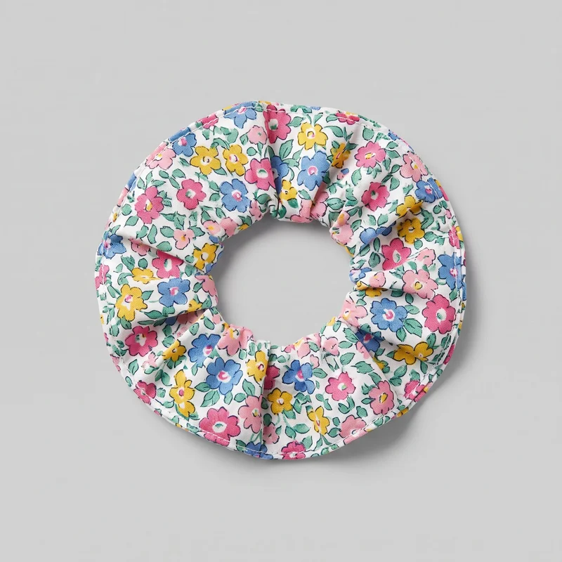 A complete fabric-covered elastic scrunchie lies flat in a top-down standalone view. Its tubular band is gathered into rounded folds around a central opening, with a continuous edge seam and an all-over floral-and-leaf print featuring outlined blossoms and foliage.