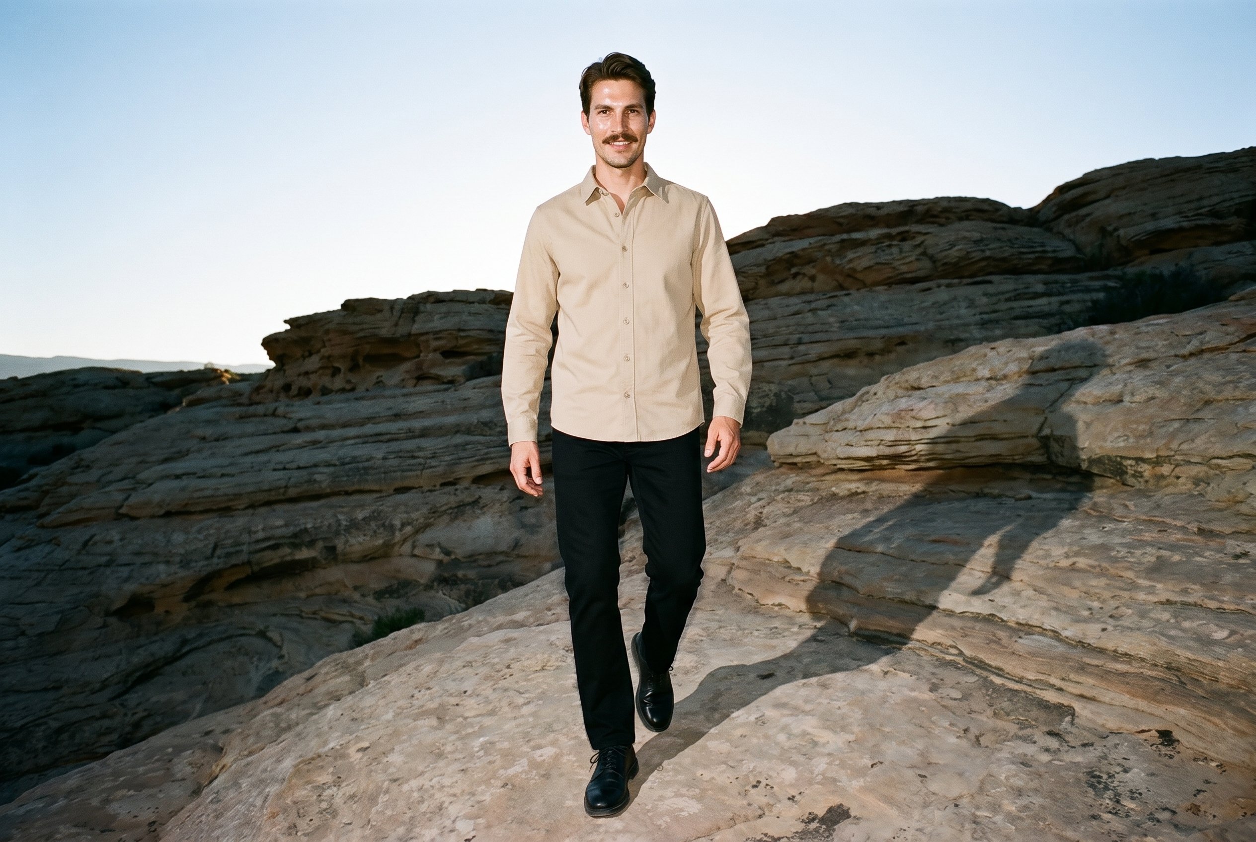 A 28-year-old Caucasian man is shown full body from the front, walking and smiling while wearing a beige twill overshirt. The Badlands serve as the backdrop.
