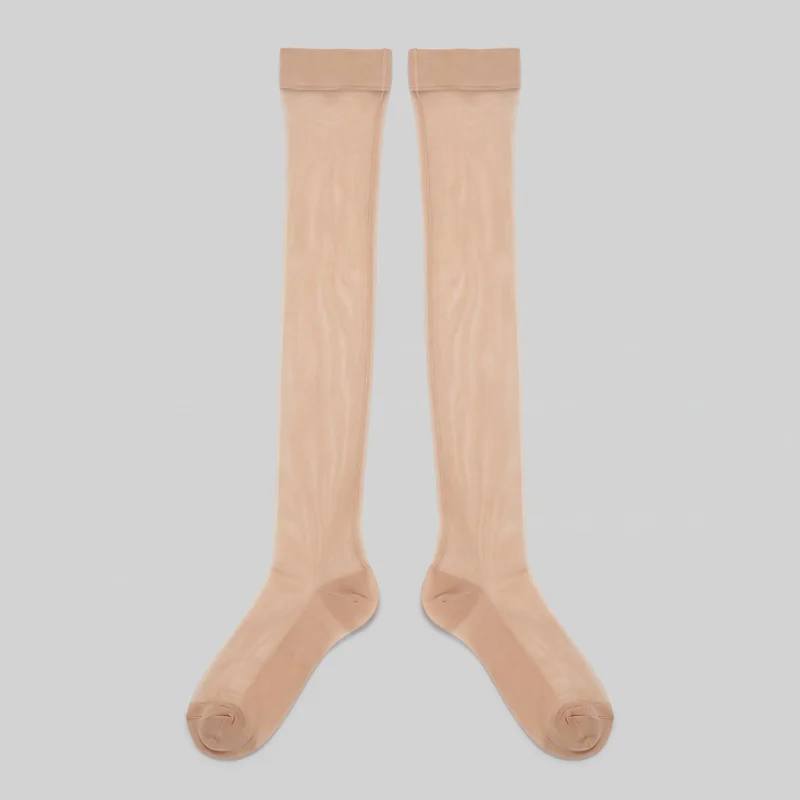 A complete pair of nude sheer thigh-high stockings is laid flat with the feet angled outward and the top bands visible. The stockings have enclosed rounded toes, reinforced-looking toe caps with curved seam lines, shaped heel areas, vertical side seams, fine sheer leg panels with subtle vertical texture, and opaque folded top bands with visible overlock stitching along the lower edges.