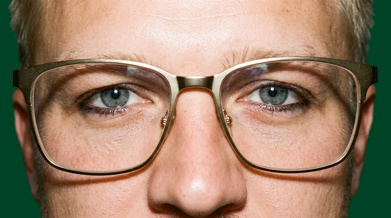 A men's gold metal optical frame shown from the front on eyes with the head tilted. The frame is presented against a flat British Racing green backdrop.