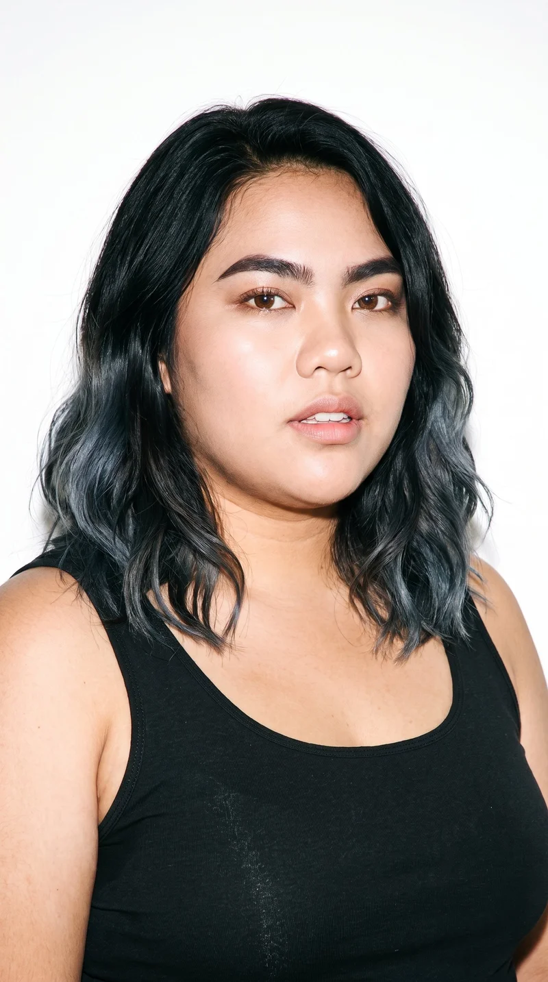 A 23-year-old Southeast Asian woman with lightly tanned skin and a warm yellow undertone. Her heart-shaped face has a wider forehead that narrows to a more pointed chin, with thick eyebrows, brown eyes, and medium-sized lips. Her black hair is styled in soft waves with gentle movement.