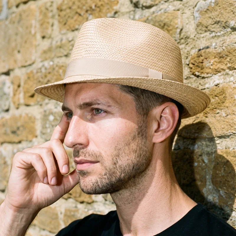 A 36-year-old Caucasian man is shown in a head-framed side view wearing a beige straw fedora, with one finger at his temple and a natural facial expression. A Cotswolds stone village backdrop is visible behind him.