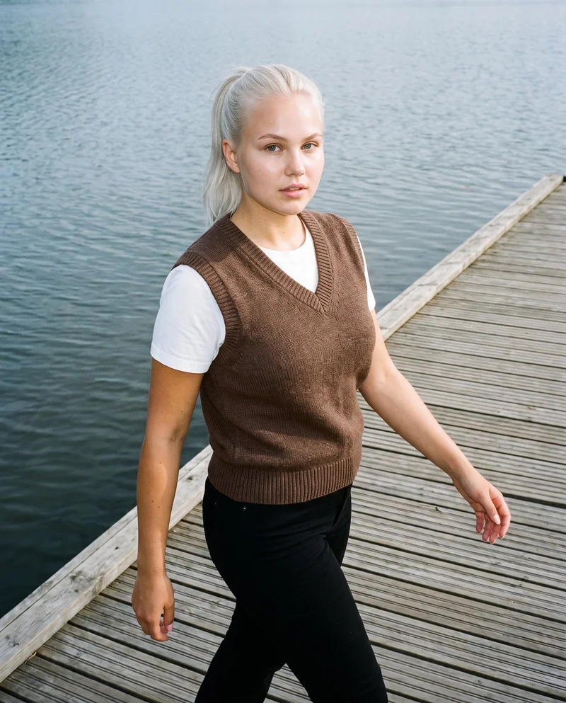 A 21-year-old Caucasian woman wearing a brown wool sweater vest is shown in a three-quarter-length, angled-front view while walking. Her expression is neutral, and a lakeside dock forms the backdrop.