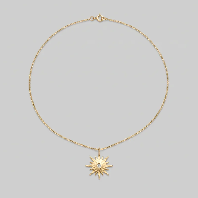 A complete women's gold link chain necklace is laid in a circular shape, with the spring-ring clasp and jump-ring hardware at the top. A centered dangling sunburst pendant hangs at the bottom, featuring irregular pointed rays and a round clear stone set in a bezel at its center.