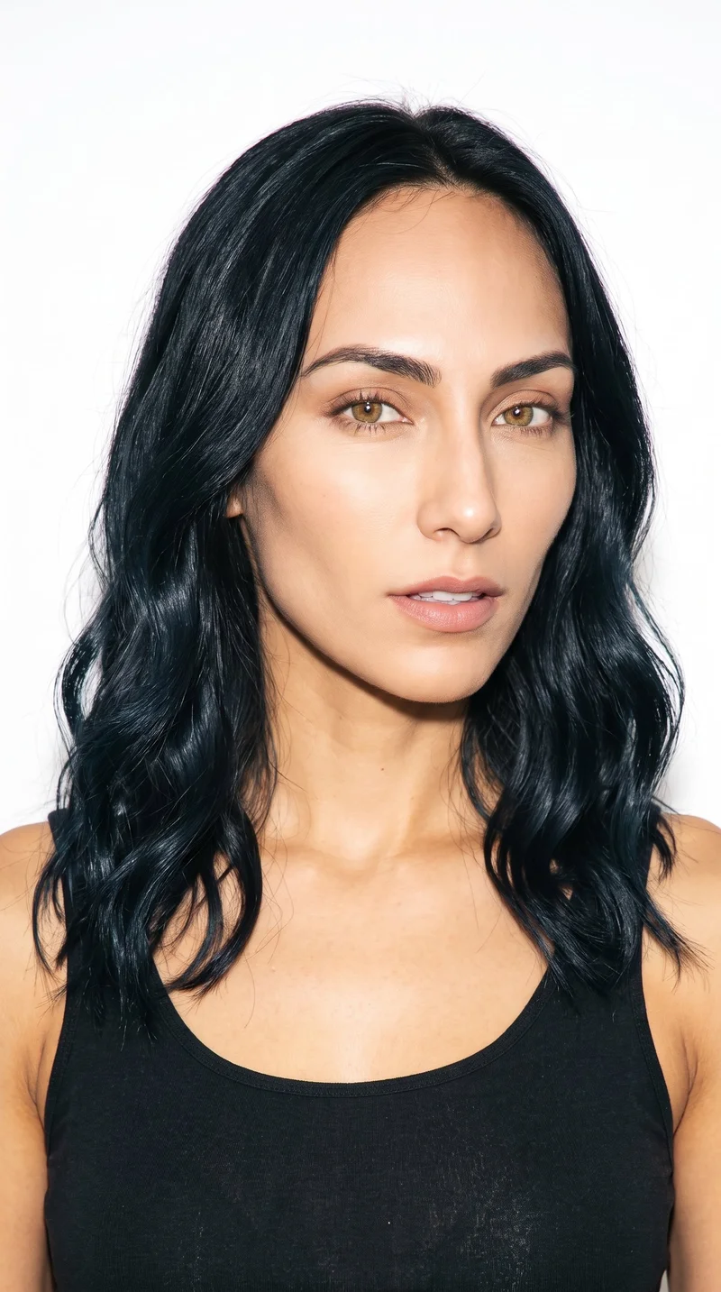 A 27-year-old Hispanic woman with warm honey-toned skin and a neutral undertone. Her face is elongated with refined features and high cheekbones. She has thick eyebrows, hazel eyes with green-gold flecks, and medium lips. Her deep black hair is styled in soft waves.