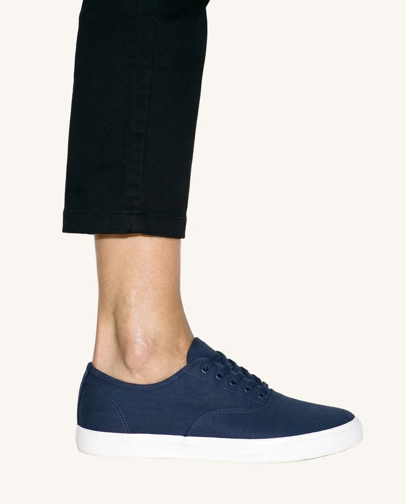 A navy canvas sneaker is shown from the side with the foot flat, framed at the ankle against a soft ivory backdrop.