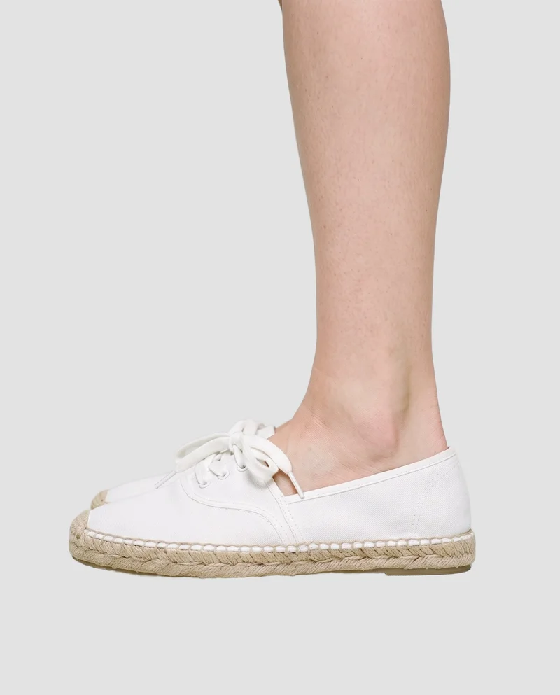A white women's espadrille is shown from the side with the foot flat, framed from the ankle downward against a flat fog-grey backdrop.