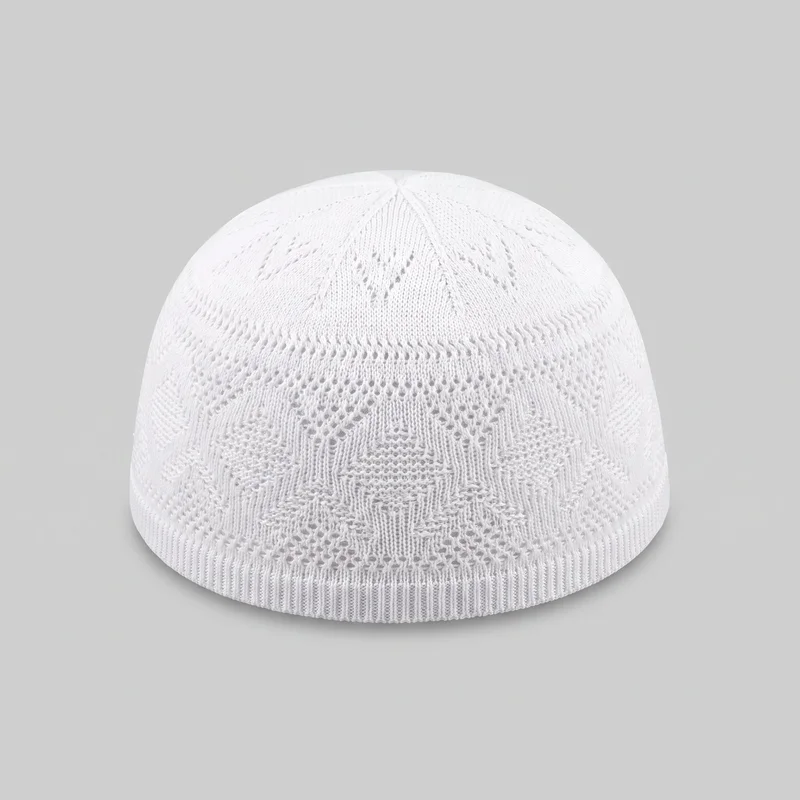 A mostly complete white knit kufi is shown alone from a slightly elevated front angle. The close-fitting rounded cap has a smooth domed crown, radial knit panels converging at the top, repeating openwork pointelle geometric motifs, denser vertical knit sections around the body, and a straight ribbed lower edge. No brim, earflaps, closures, logos, patches, prints, embroidery, or embellishments are visible.