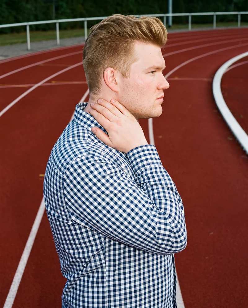 A 23-year-old Caucasian male model wears a navy gingham button-down shirt in an upper-body side view, with one hand resting on his neck and a serious expression. A running track forms the backdrop.