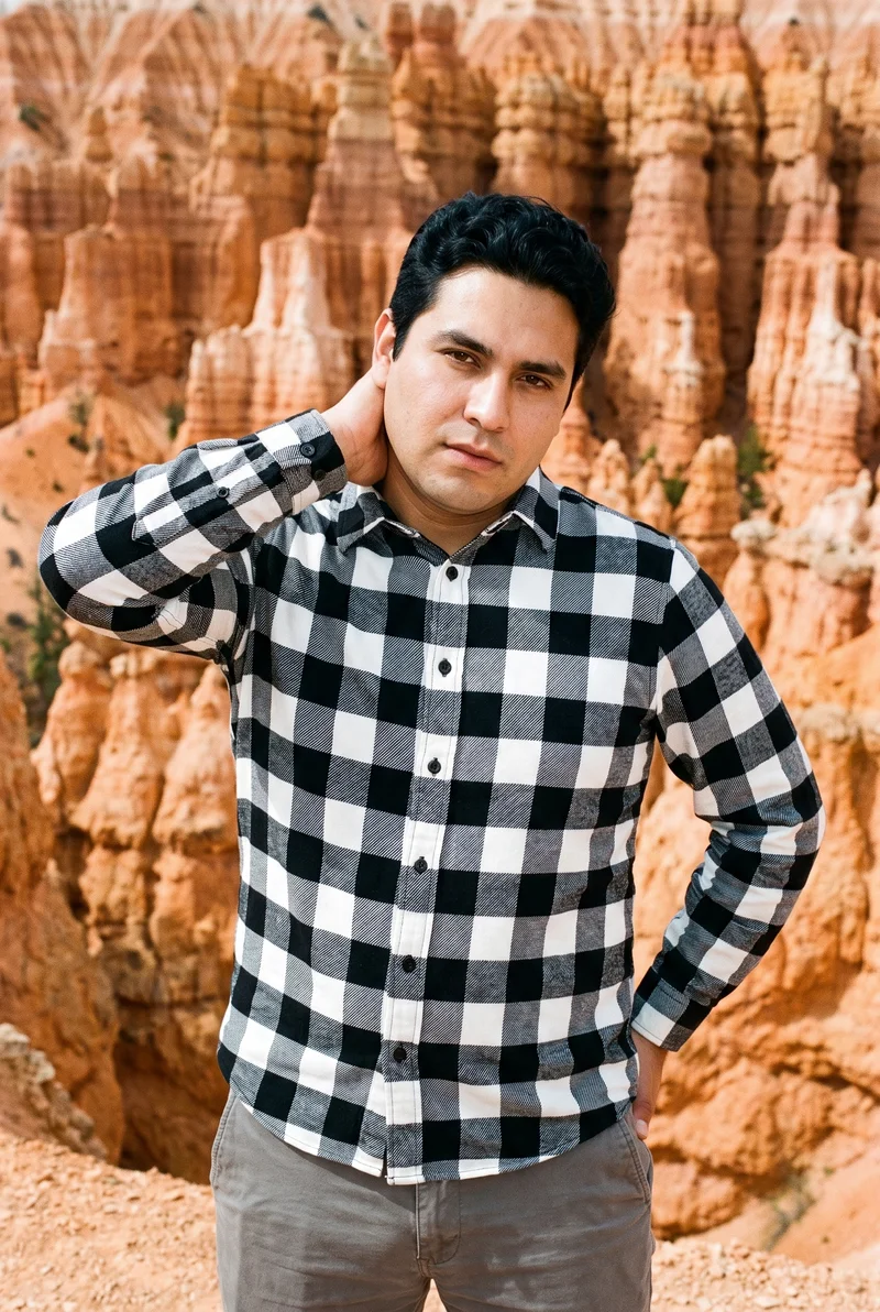 A 31-year-old Hispanic male model wears a black and white buffalo-check flannel shirt in an upper-body view from a front three-quarter angle, with one hand behind his neck and a confident expression. Bryce Canyon hoodoos form the backdrop.