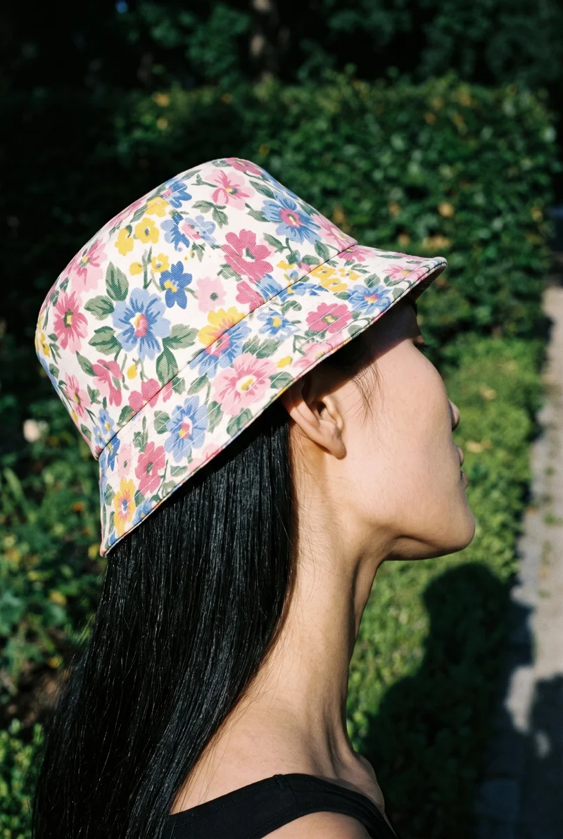 A 21-year-old Korean woman wears a women's floral print bucket hat, shown in a head crop from an angled rear view with her head tilted. A park pathway forms the backdrop.