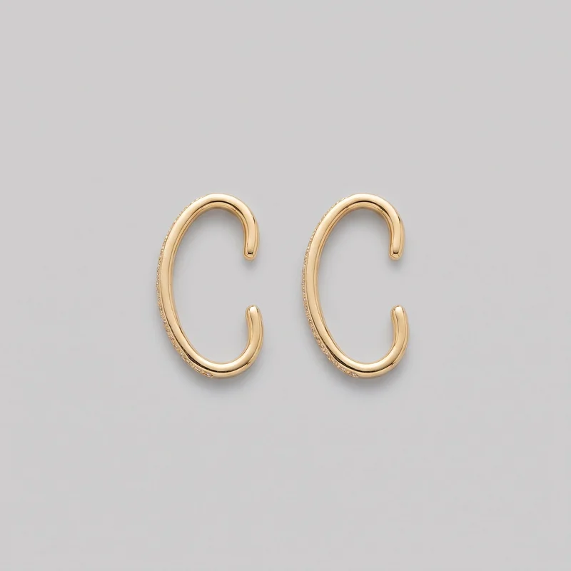 A pair of open C-shaped hoop ear cuffs is shown as a standalone product. Each polished gold-tone cuff has an elongated oval curve, a side opening with rounded capped ends, raised inner and outer contours, fine groove lines, and a pavé-like strip of clear stones along the outer edge.